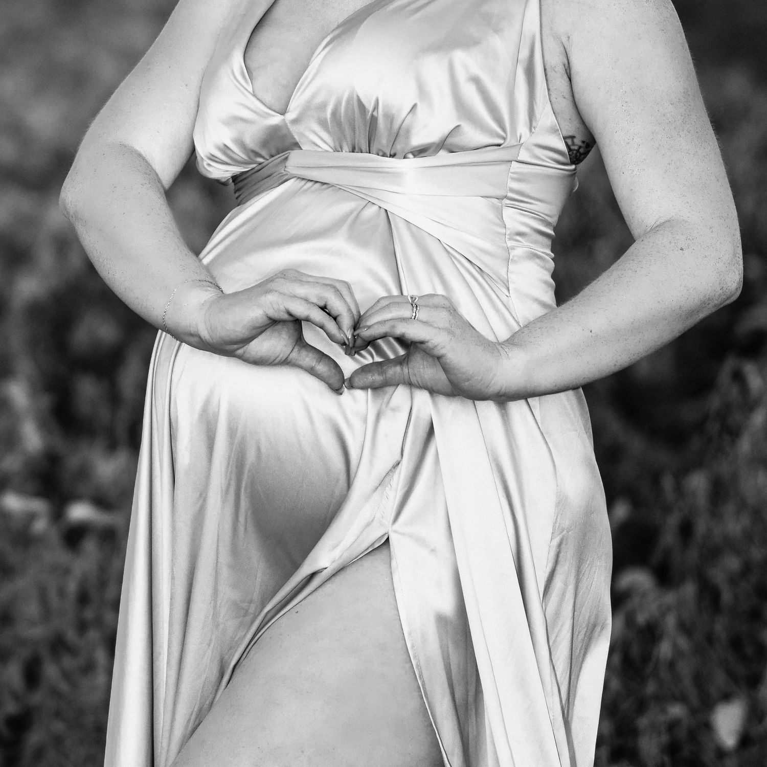 Pregnant woman in elegant satin gown making heart shape with hands over baby bump, black and white maternity photo.