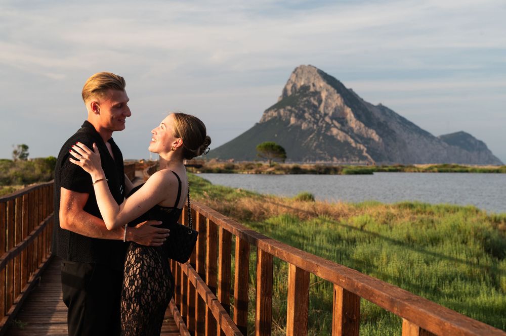 Couple embracing on wooden boardwalk with dramatic mountain and wetland landscape backdrop.