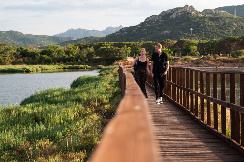Couple walking along wooden boardwalk with mountain ranges and wetlands in background.