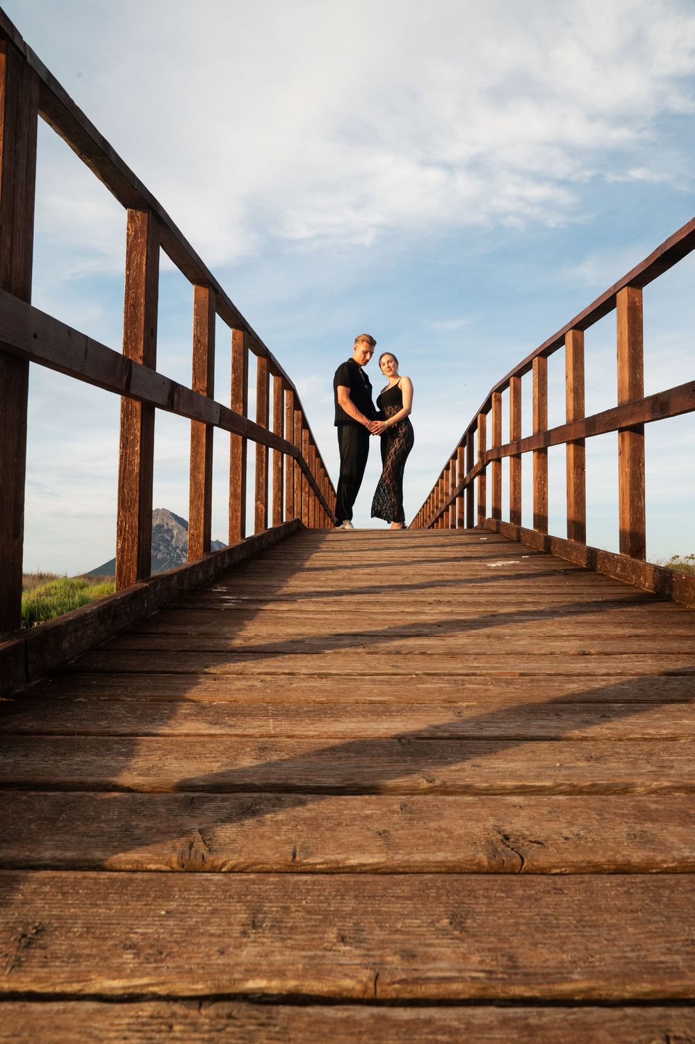 A couple stands silhouetted against the sky on a rustic wooden boardwalk.
