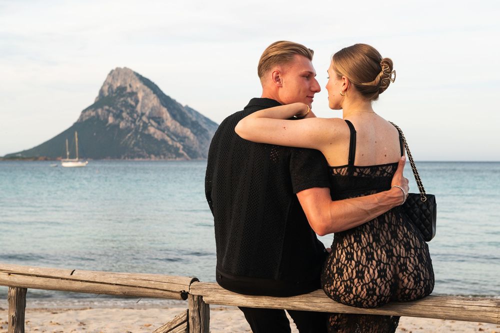Couple sitting together on a beach with a mountain backdrop and sailboat in the distance.