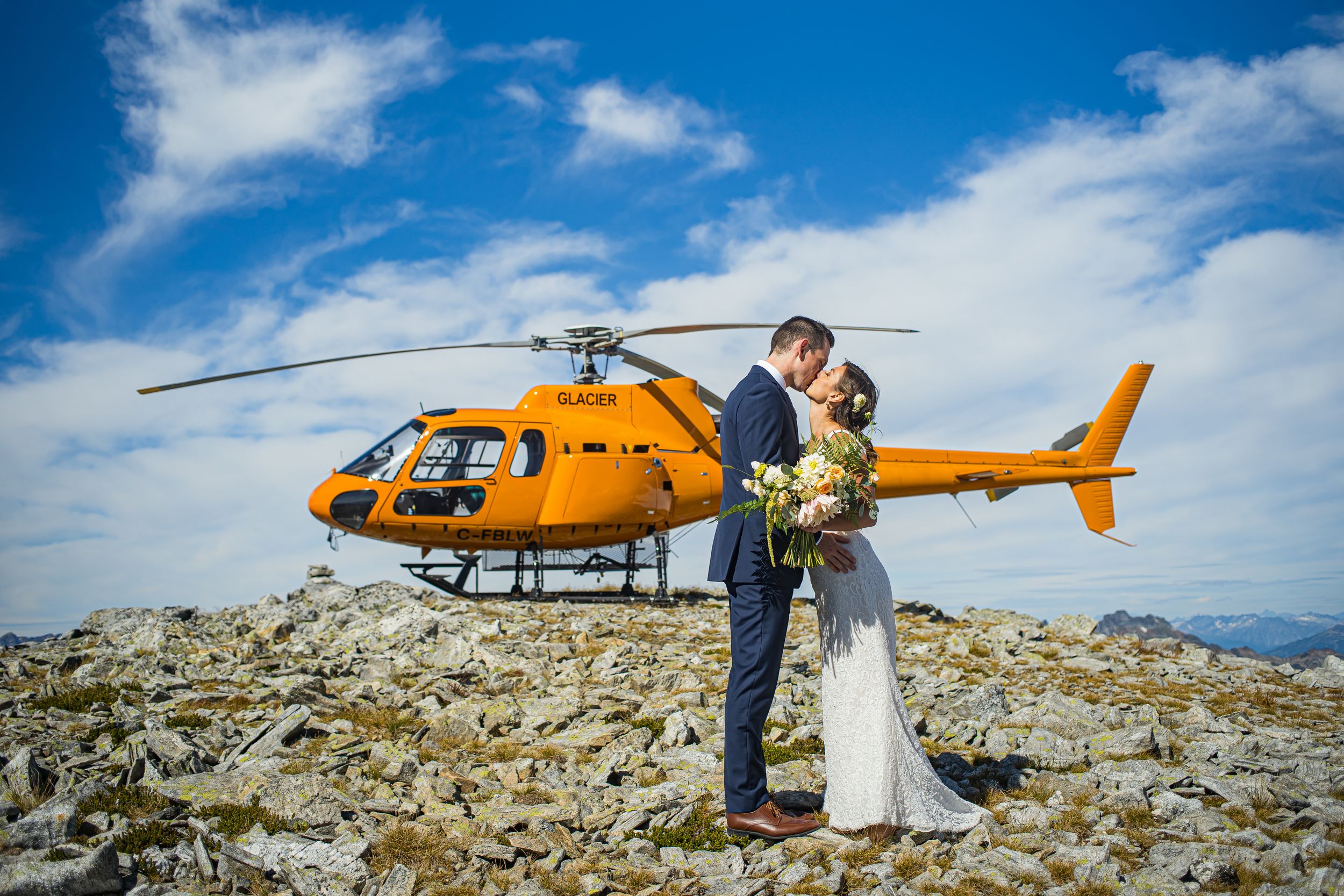 Bride and groom standing beside helicopter on a mountain ridge overlooking Revelstoke — professional wedding photography.