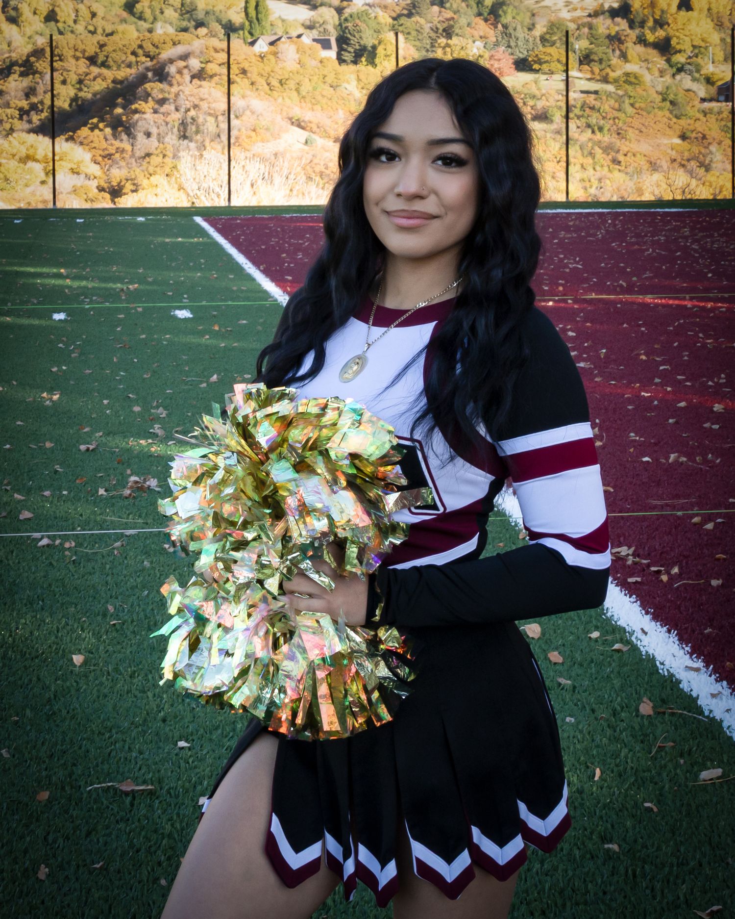 A cheerleader in black and white uniform holds gold pom-poms while posing on a sports field with mountains in background.