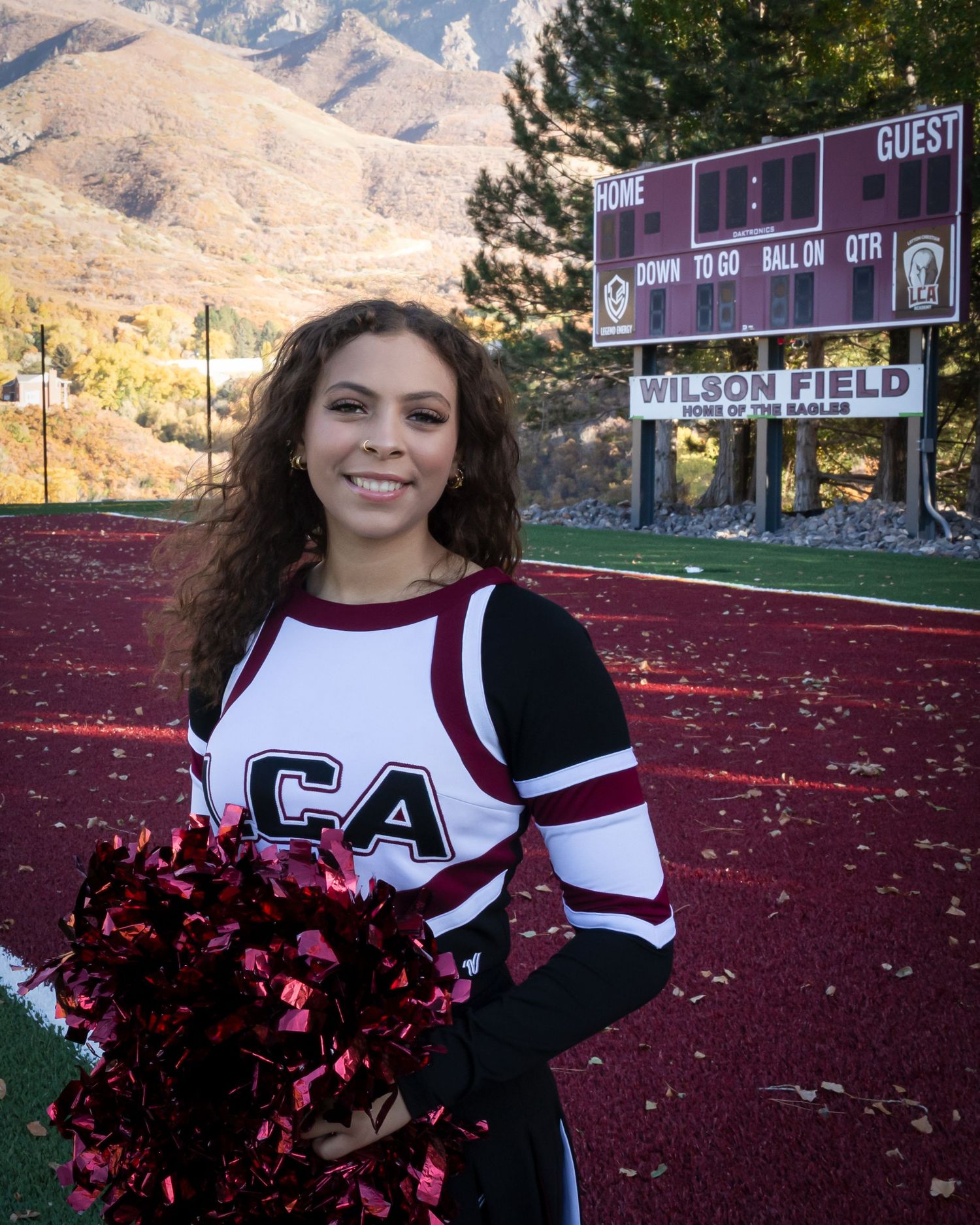 A cheerleader in maroon and white uniform poses in front of Wilson Field with mountains visible in the background.