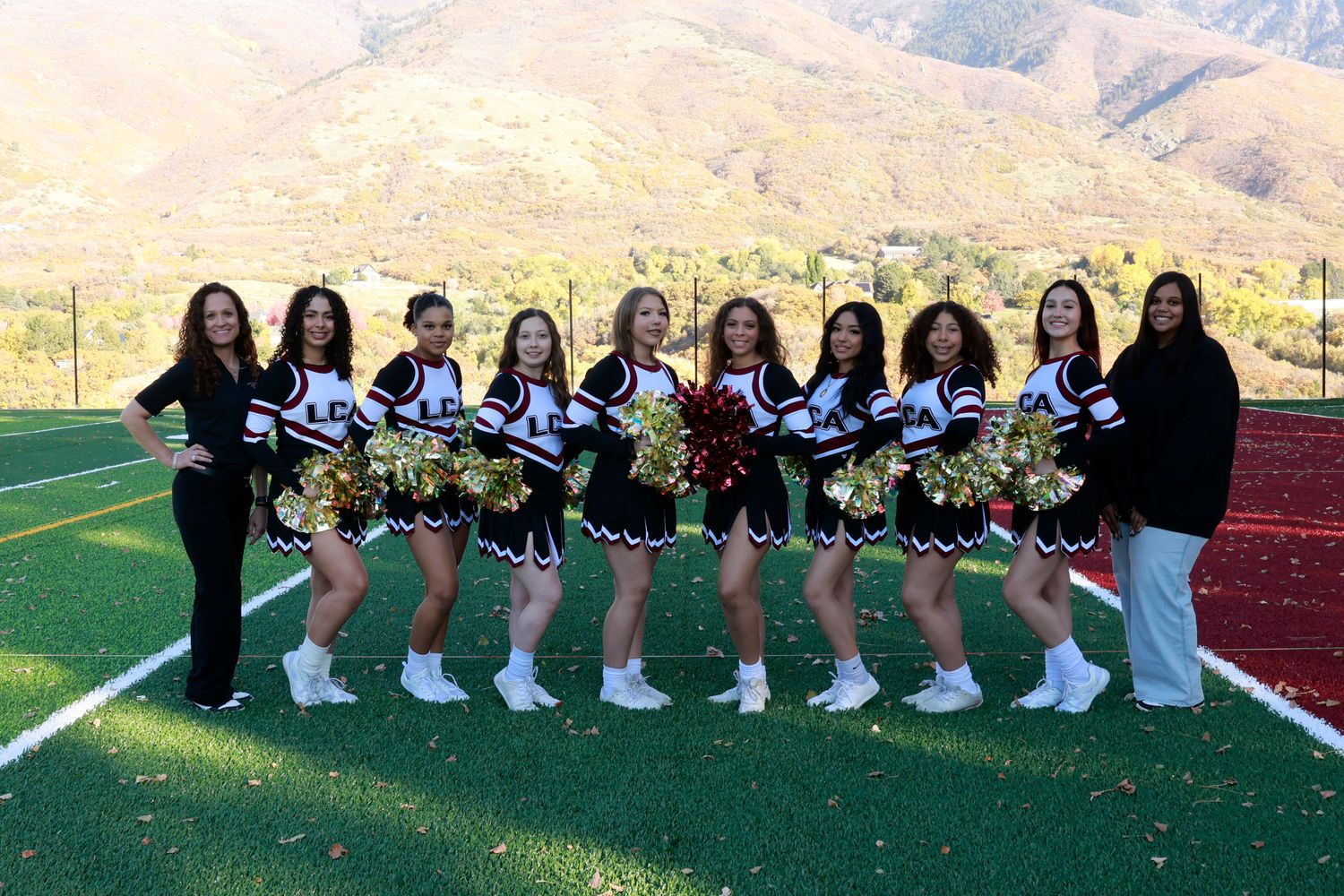 A group of cheerleaders in blue and white uniforms pose together on a red track with mountains in the background.