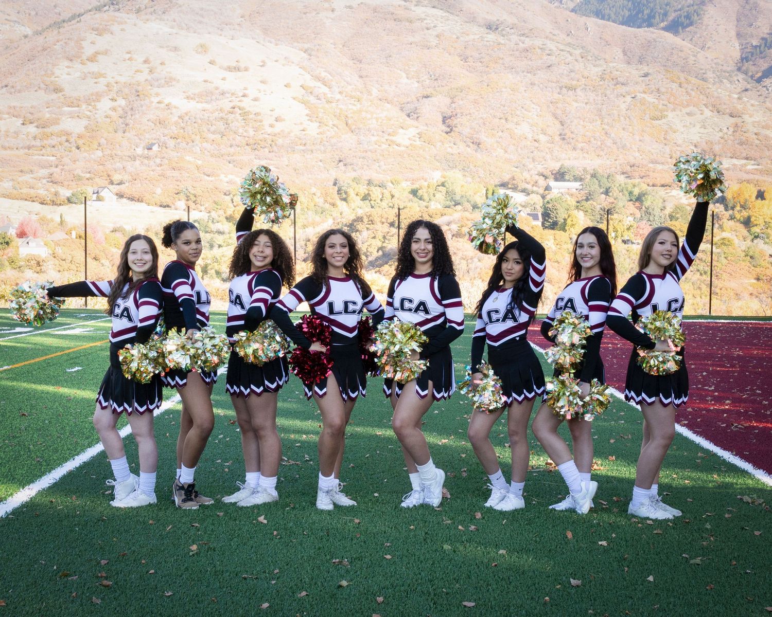 Cheerleaders in black and white uniforms pose with pom poms on a sports field with mountains in the background.