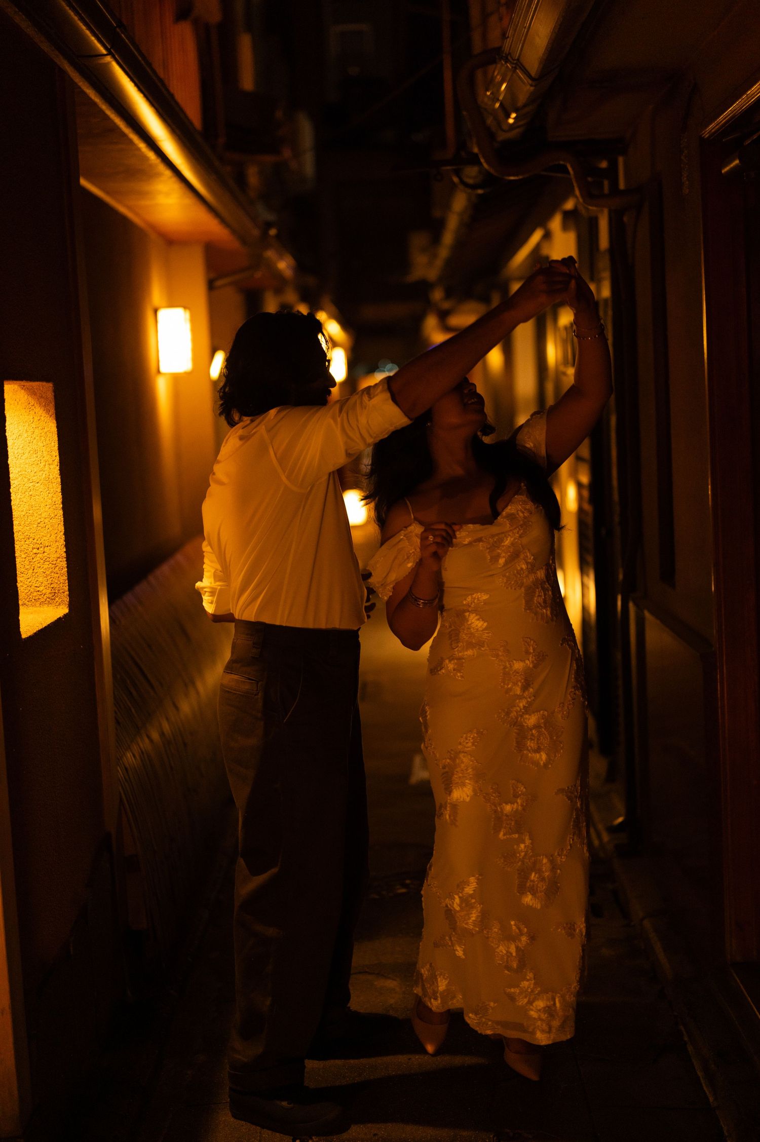 Couple dancing playfully in a golden-lit narrow Japanese alley at night.