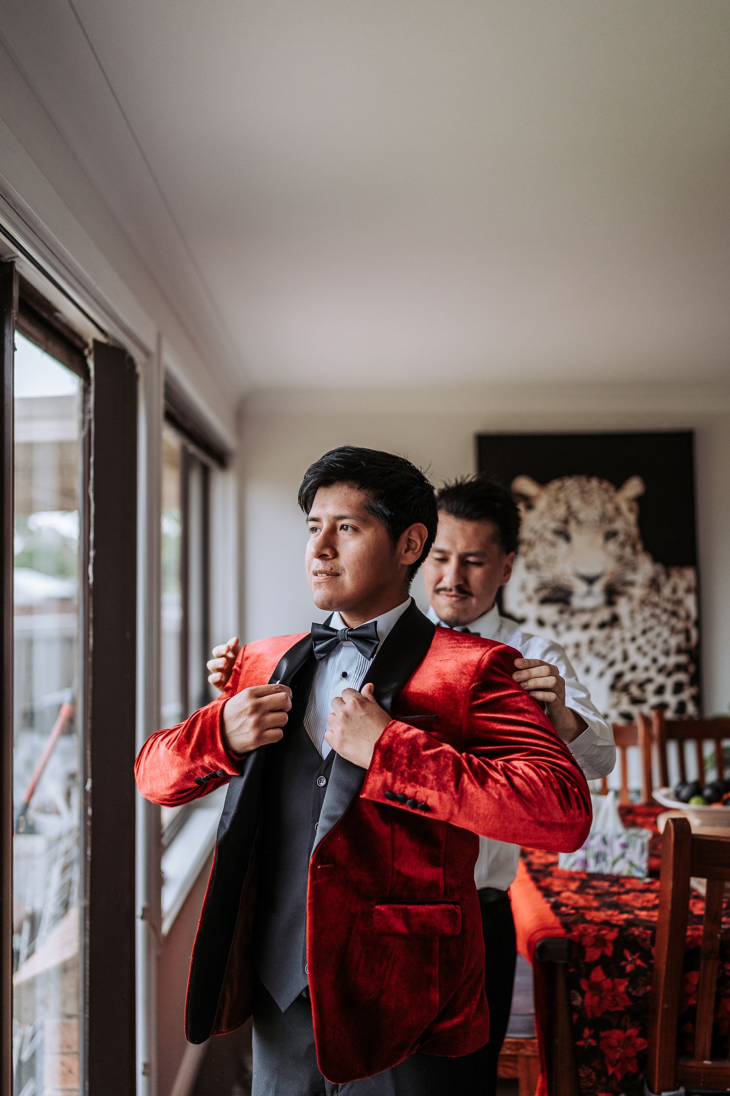 Two people in formal attire help each other adjust their ties while standing near windows in a well-lit room.