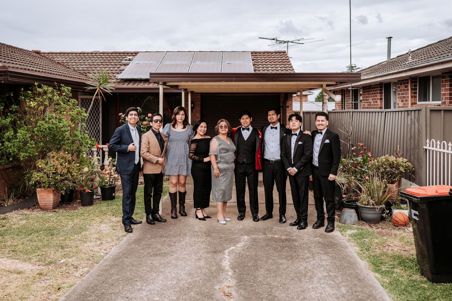 A group of formally dressed people gather outside a suburban home with brick walls and potted plants.