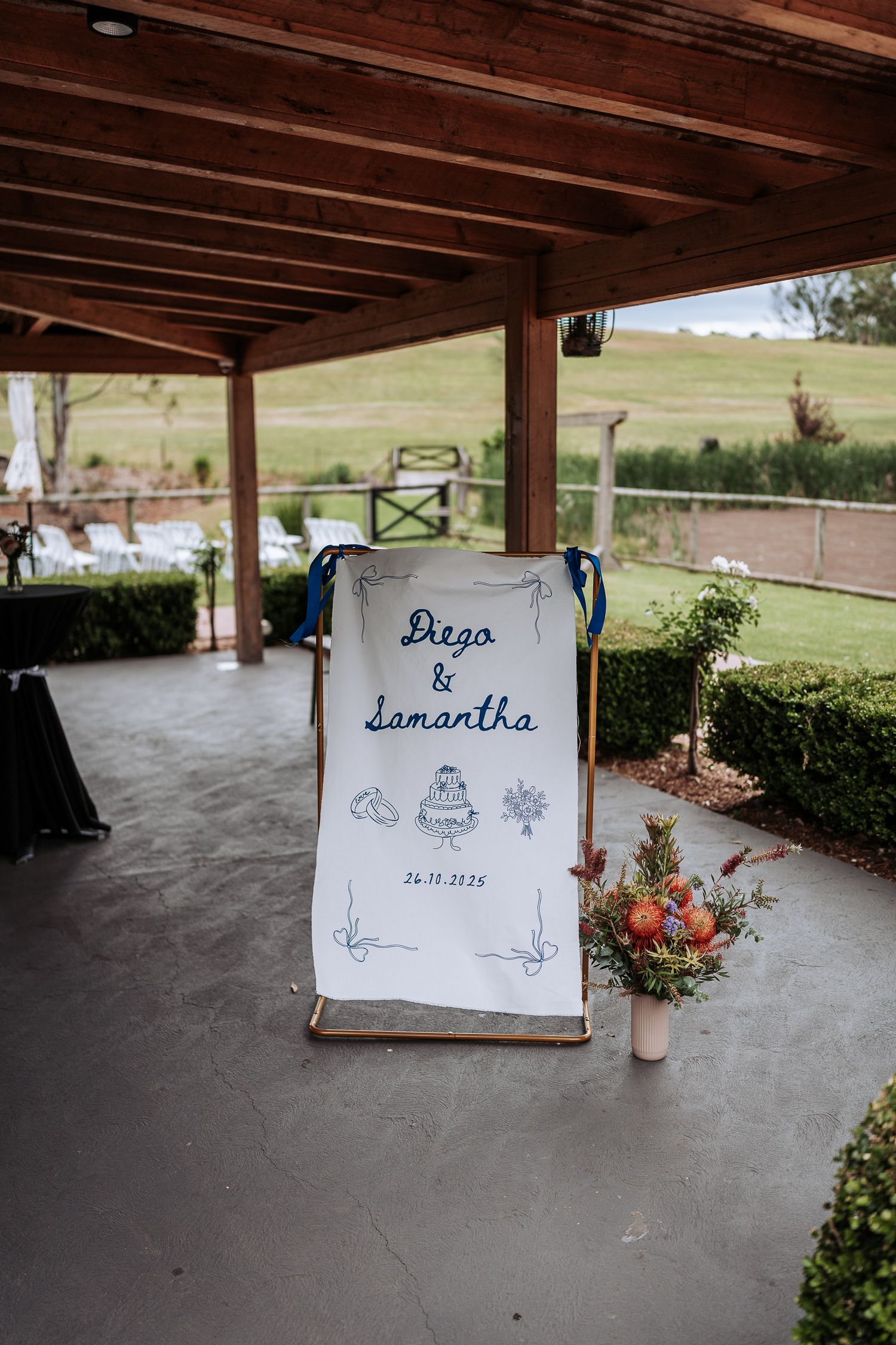 Rustic outdoor wedding venue displays personalized signs and floral arrangements under a wooden pavilion.