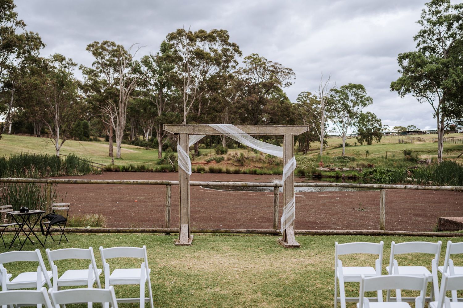 An outdoor wedding ceremony setup with white chairs and a wooden arch against a rural Australian landscape.