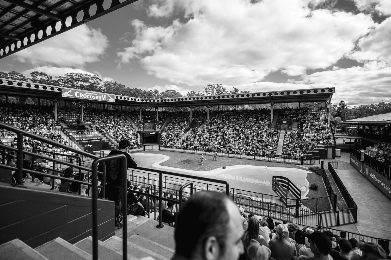 Black and white panoramic view of a packed grandstand at a historic race track with dramatic cloudy sky overhead.