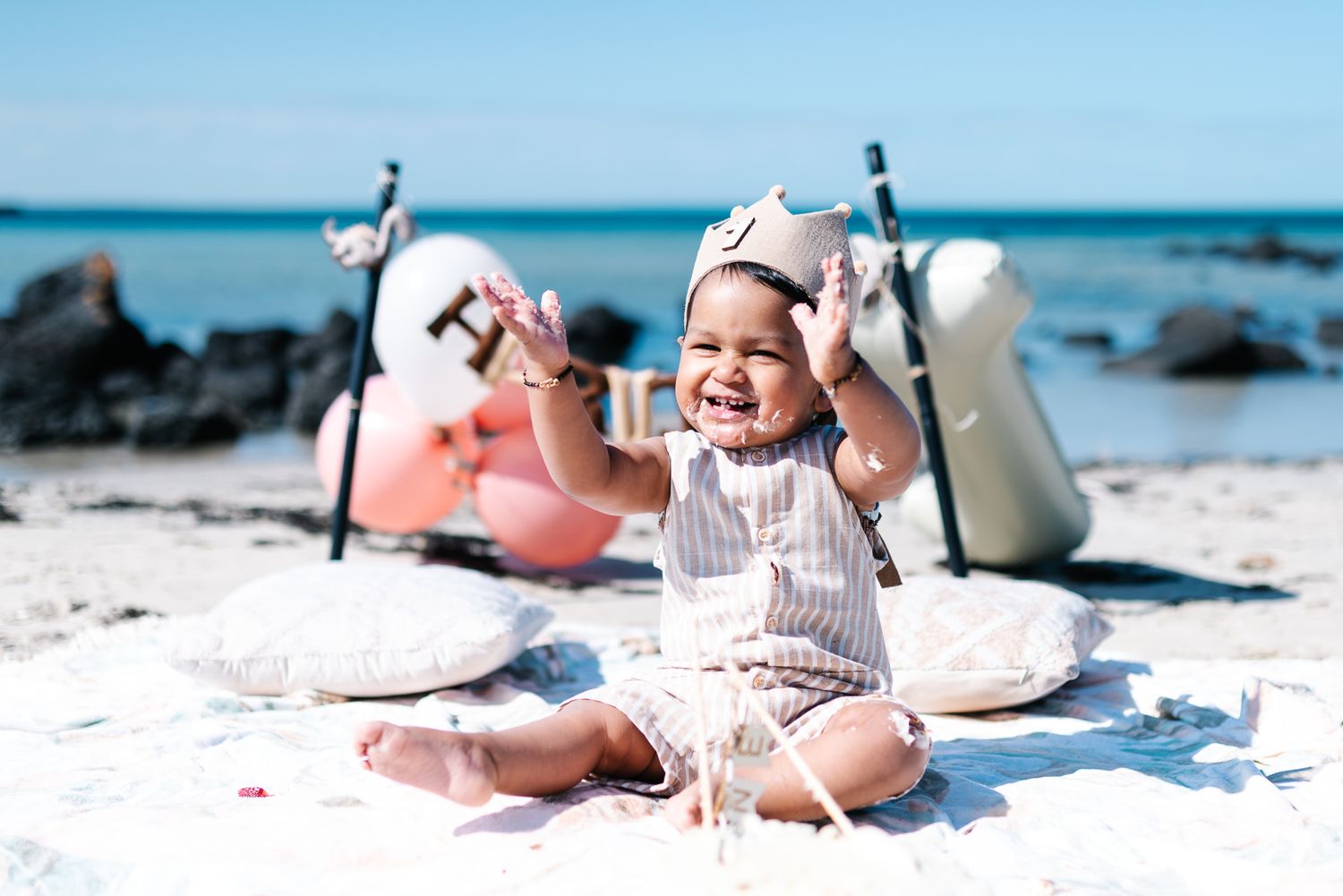 Baby lying on white sand beach with seashells and turquoise ocean water in background.