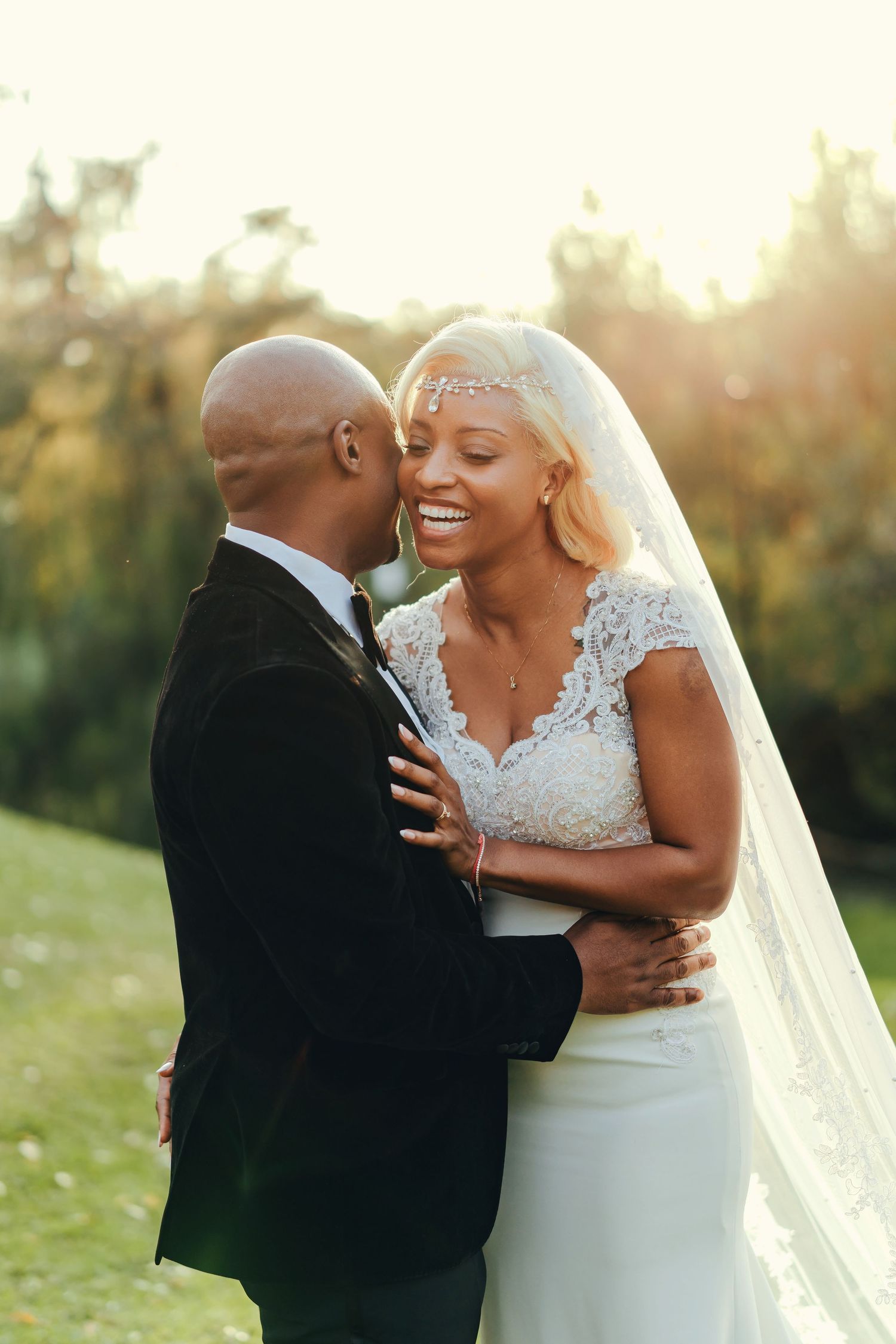 A joyful wedding couple embraces outdoors during golden hour sunset, with the bride wearing a lace cap sleeve gown and veil.