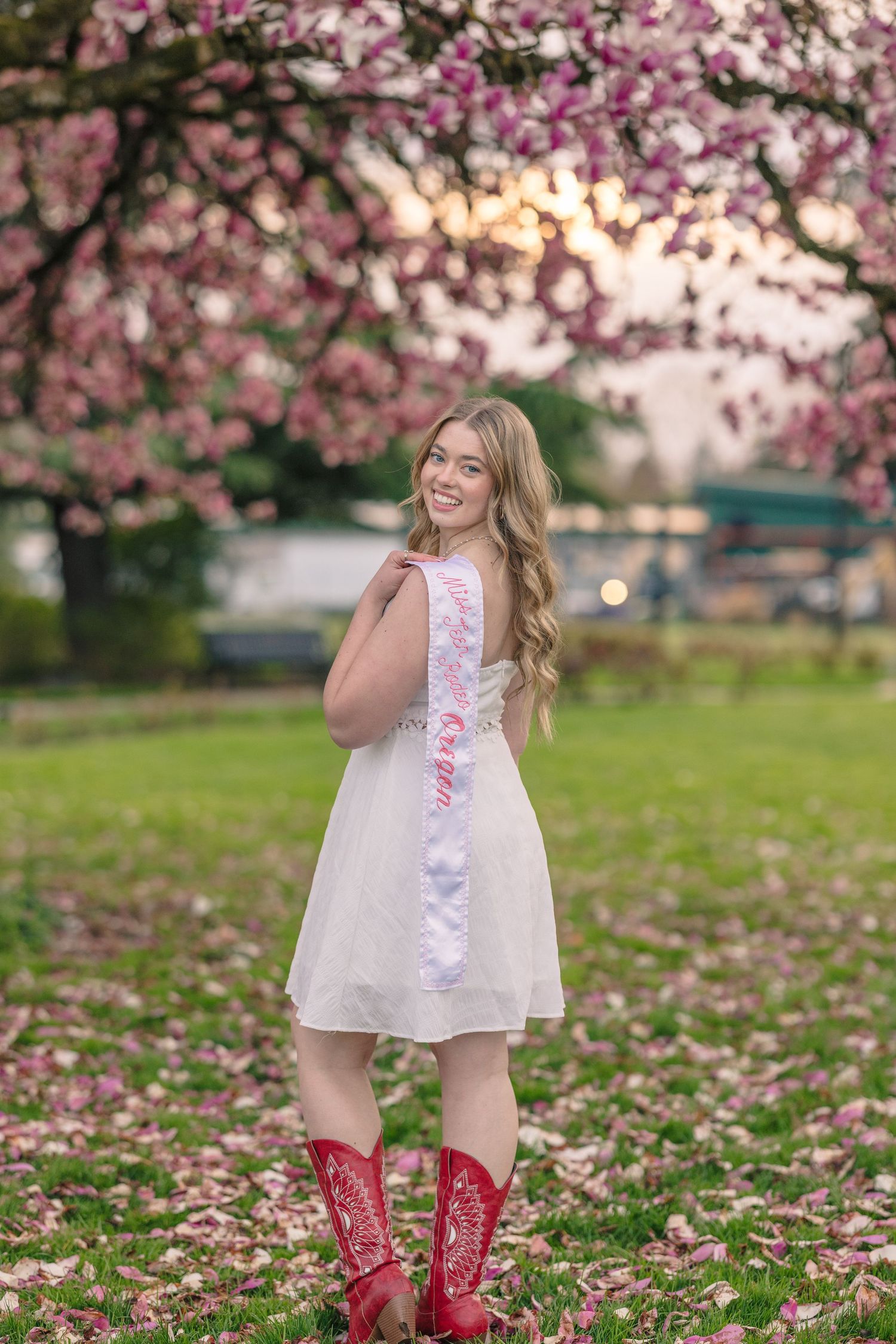 Child wearing a white dress and red cowboy boots under blooming cherry blossoms.