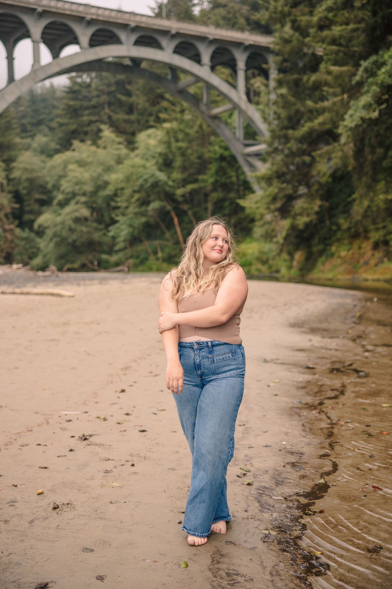 Person sitting by riverbank under arched bridge on sandy shore.