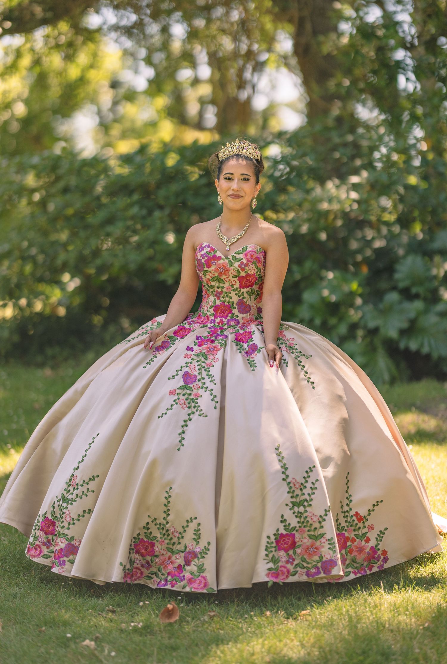 Beautiful quinceañera dress with pink floral embroidery photographed in outdoor garden setting.