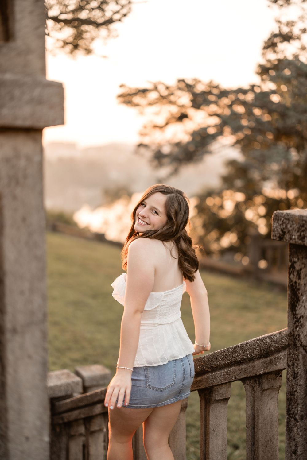 A person in a white top and denim skirt leans on a rustic wooden fence at sunset in a pastoral setting.
