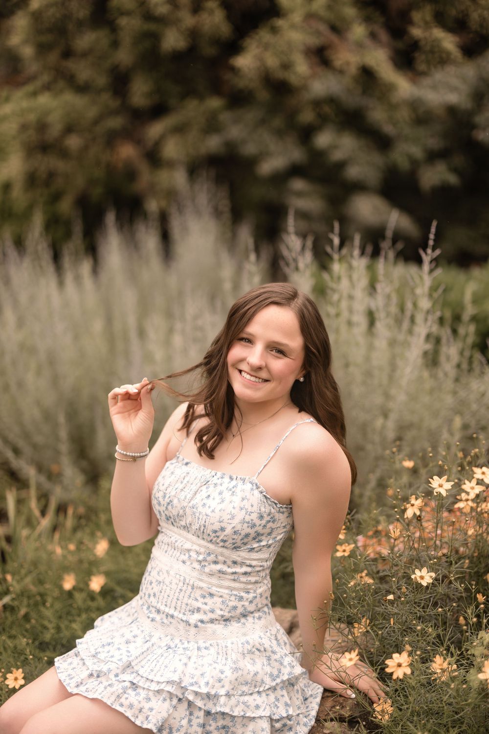 A person in a white floral dress sits in a meadow with wildflowers and tall grass on a sunny summer day.