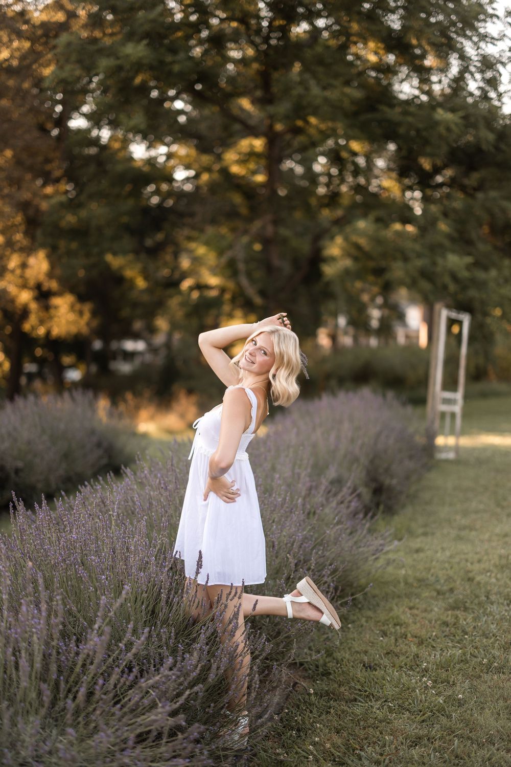 A person in a white summer dress poses playfully in a garden with blooming lavender bushes at golden hour.