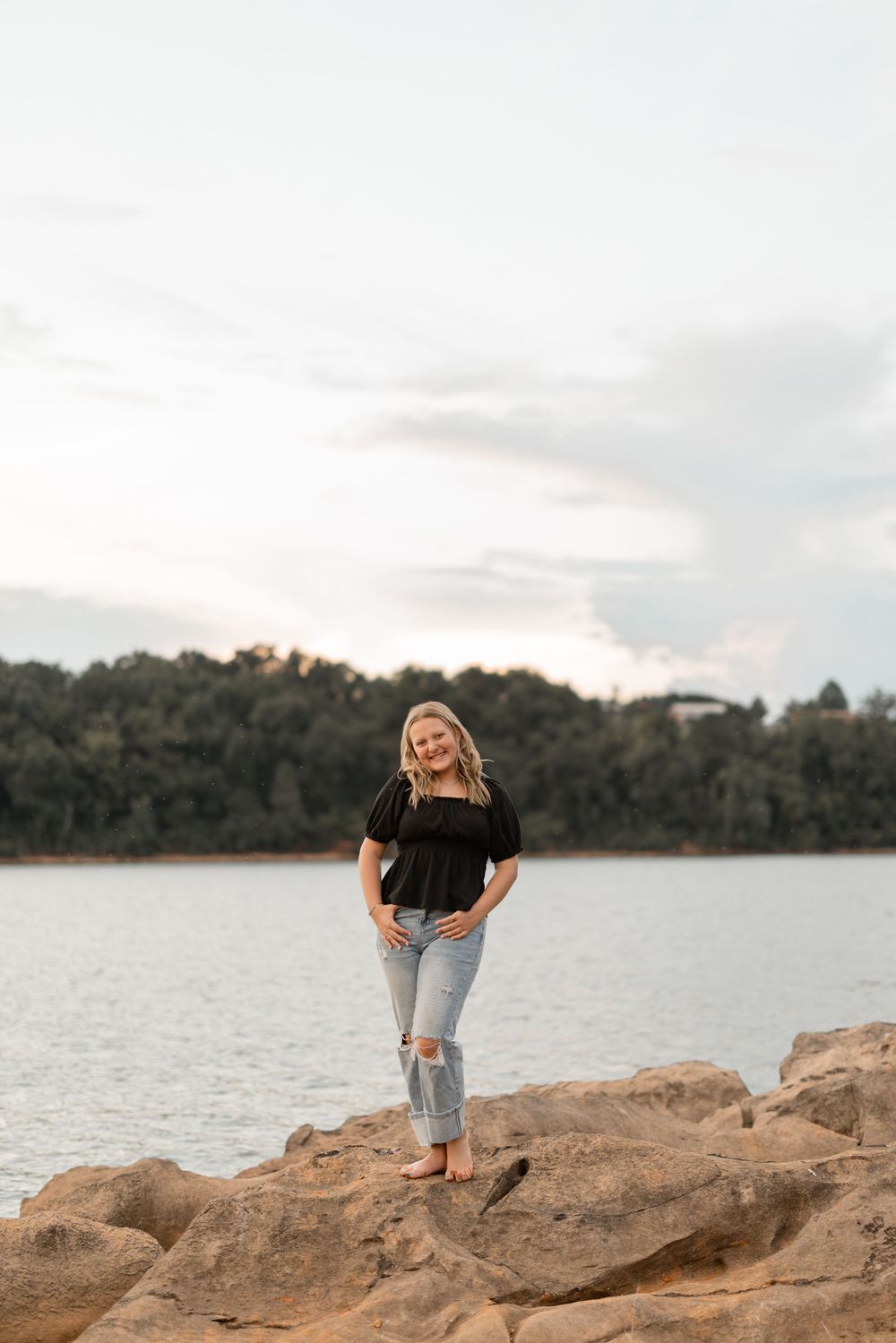 Person in casual black top and ripped jeans poses on rocky shoreline by tranquil lake at sunset.