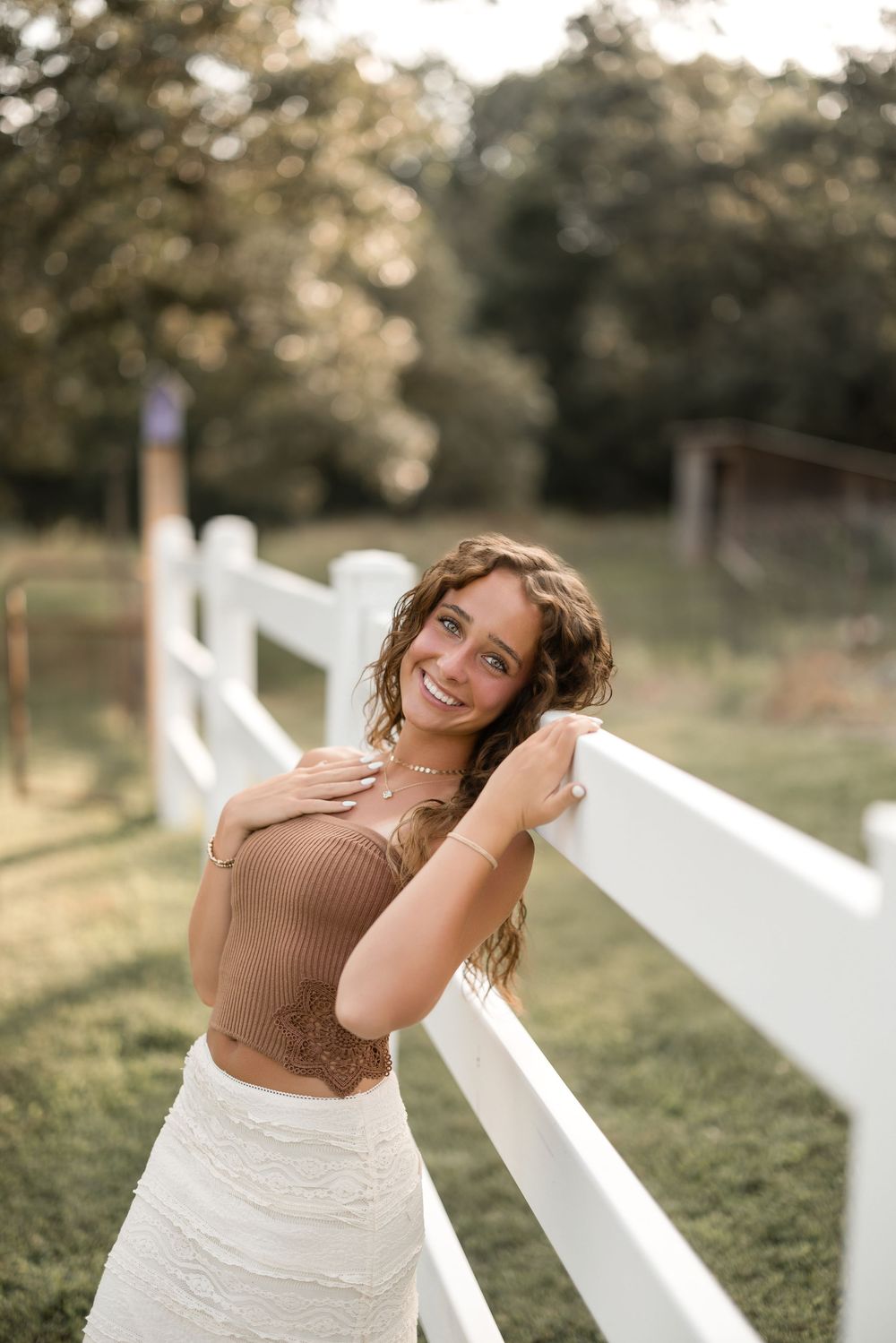 A person in a brown sweater and white skirt poses joyfully by a white fence in a sunlit outdoor setting.