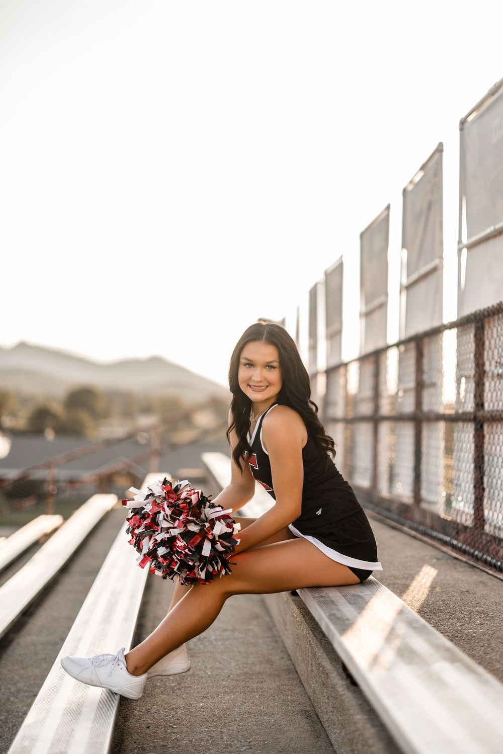 Cheerleader in black uniform sits on bleachers holding red and white pom poms during golden hour photoshoot.
