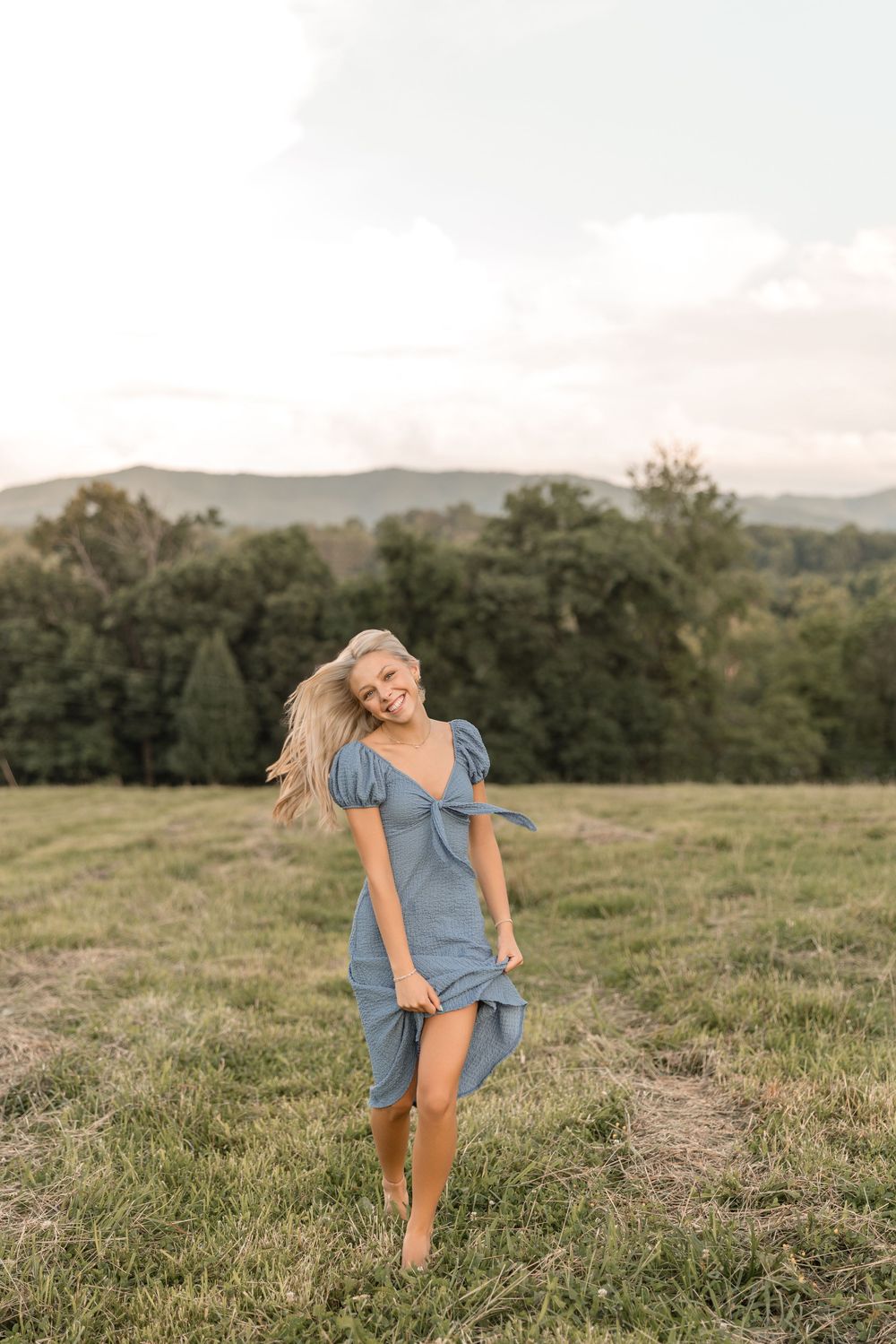 A woman in a blue dress dances joyfully in a grassy meadow with mountains in the background.