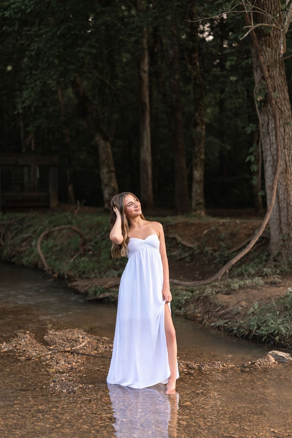 A figure in a flowing white dress walks through a wooded forest path during golden hour.