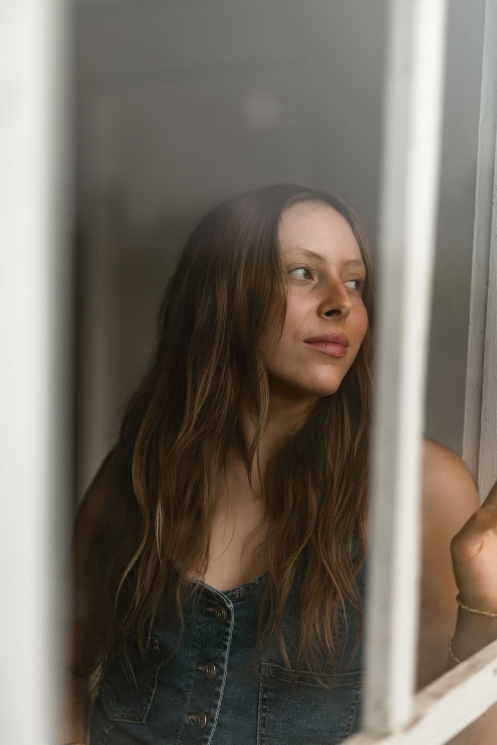 Woman with long brown hair in denim jacket gazes through white bars in artistic portrait.
