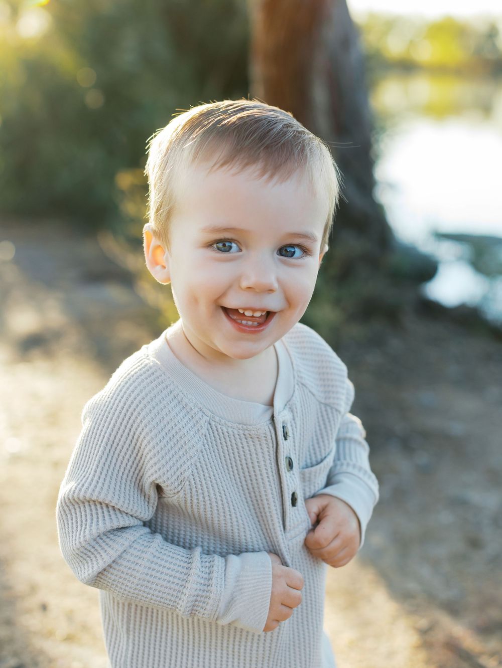 A small child wearing a cozy gray knit sweater smiles brightly in warm outdoor lighting.