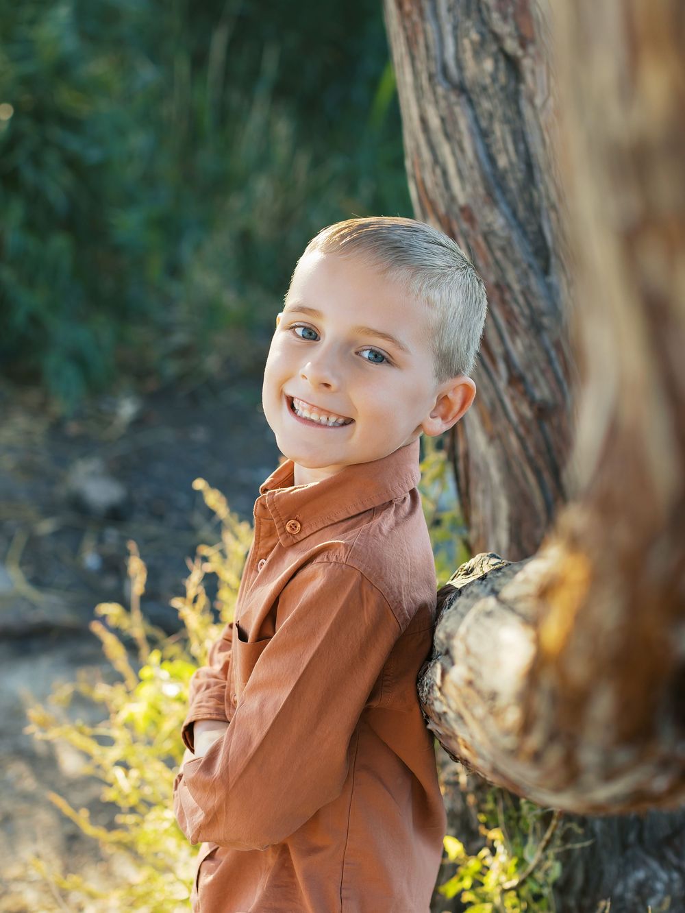 Young child in brown jacket poses playfully against natural outdoor backdrop with yellow foliage and trees.