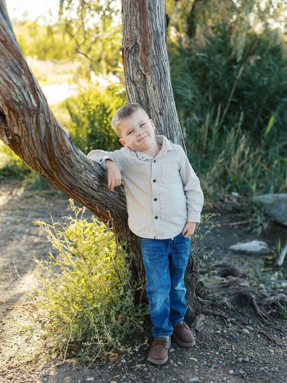 Young child in blue jeans and white sweater stands by tree trunk in outdoor nature setting at golden hour.