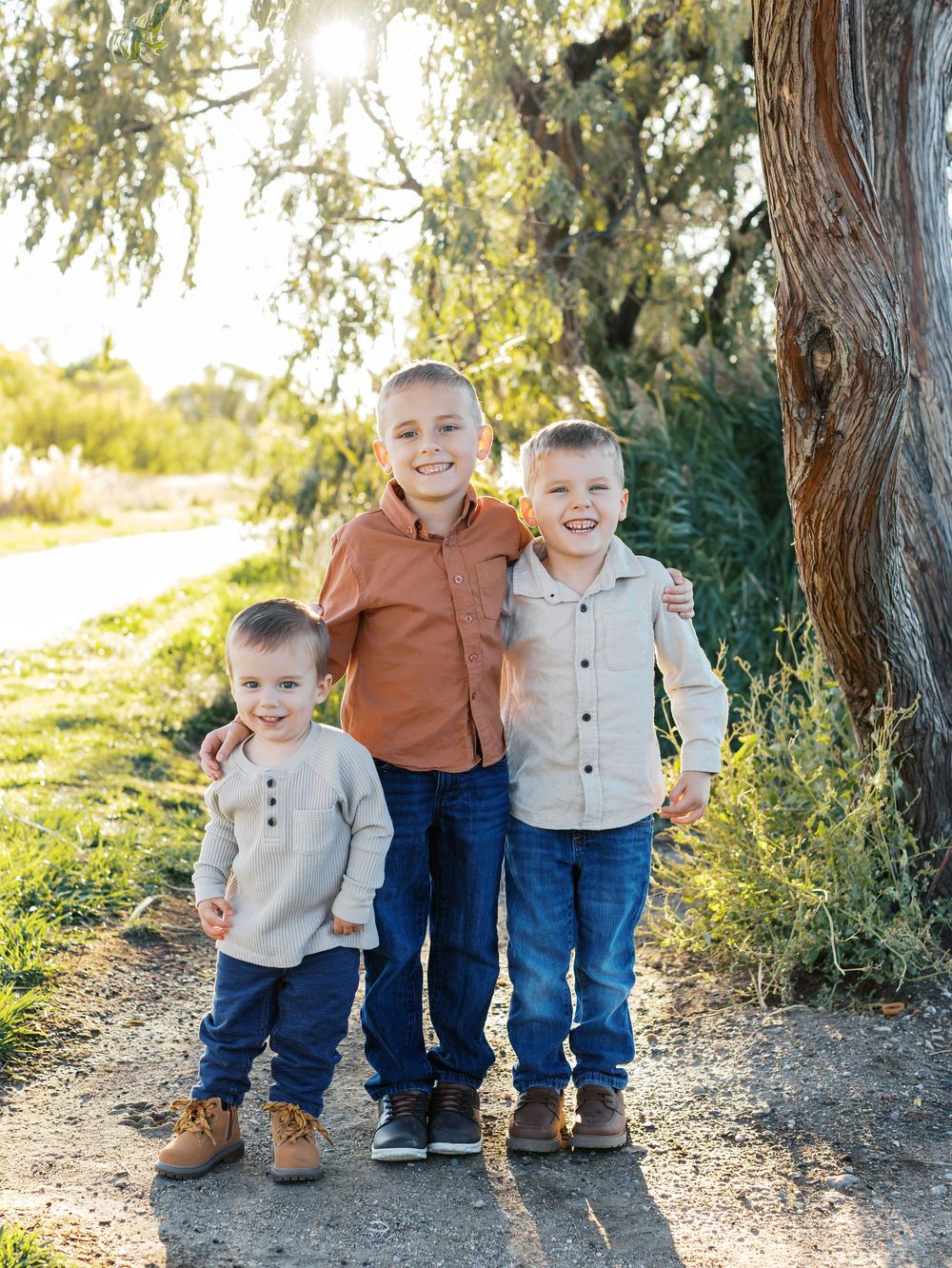 Three young children stand together on a dirt path near trees during golden hour.