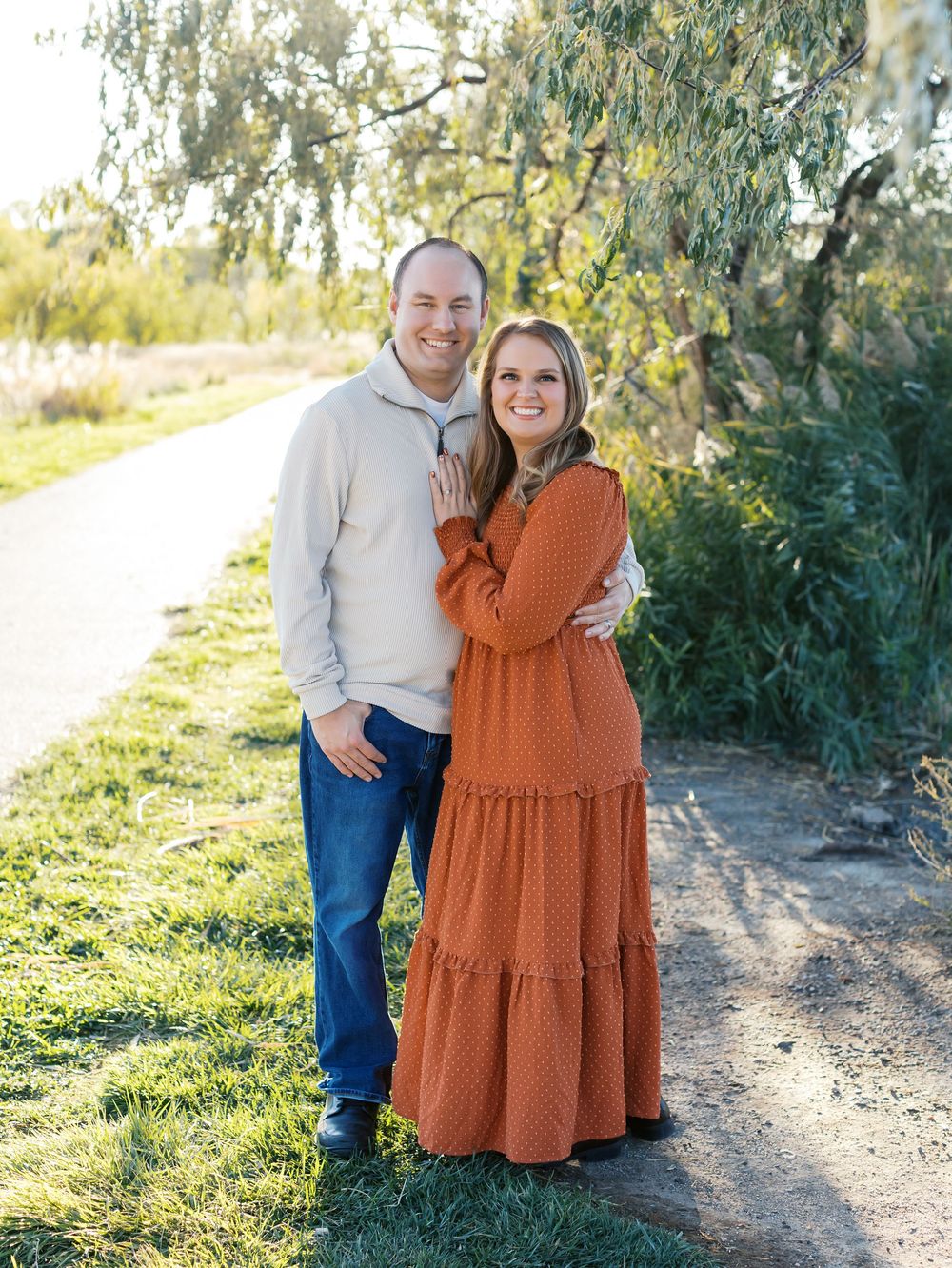 Couple in casual sweaters embraces on a sunlit nature path with trees in the background.