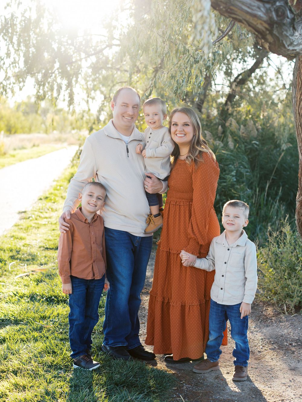 A family poses together under a willow tree at sunset wearing coordinated fall outfits in rust and cream colors.