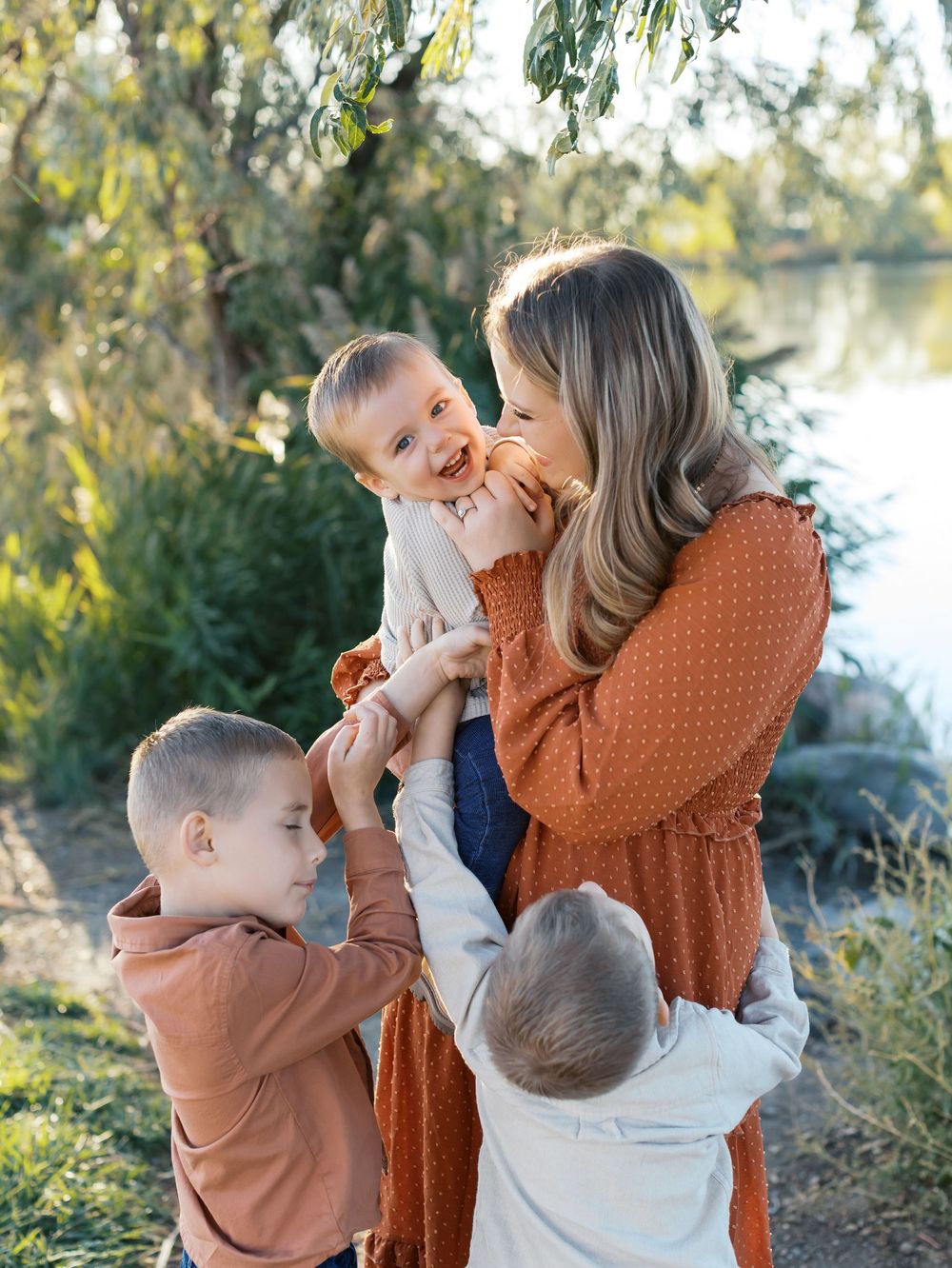 Family shares tender moments together outdoors with sunlit trees in background.