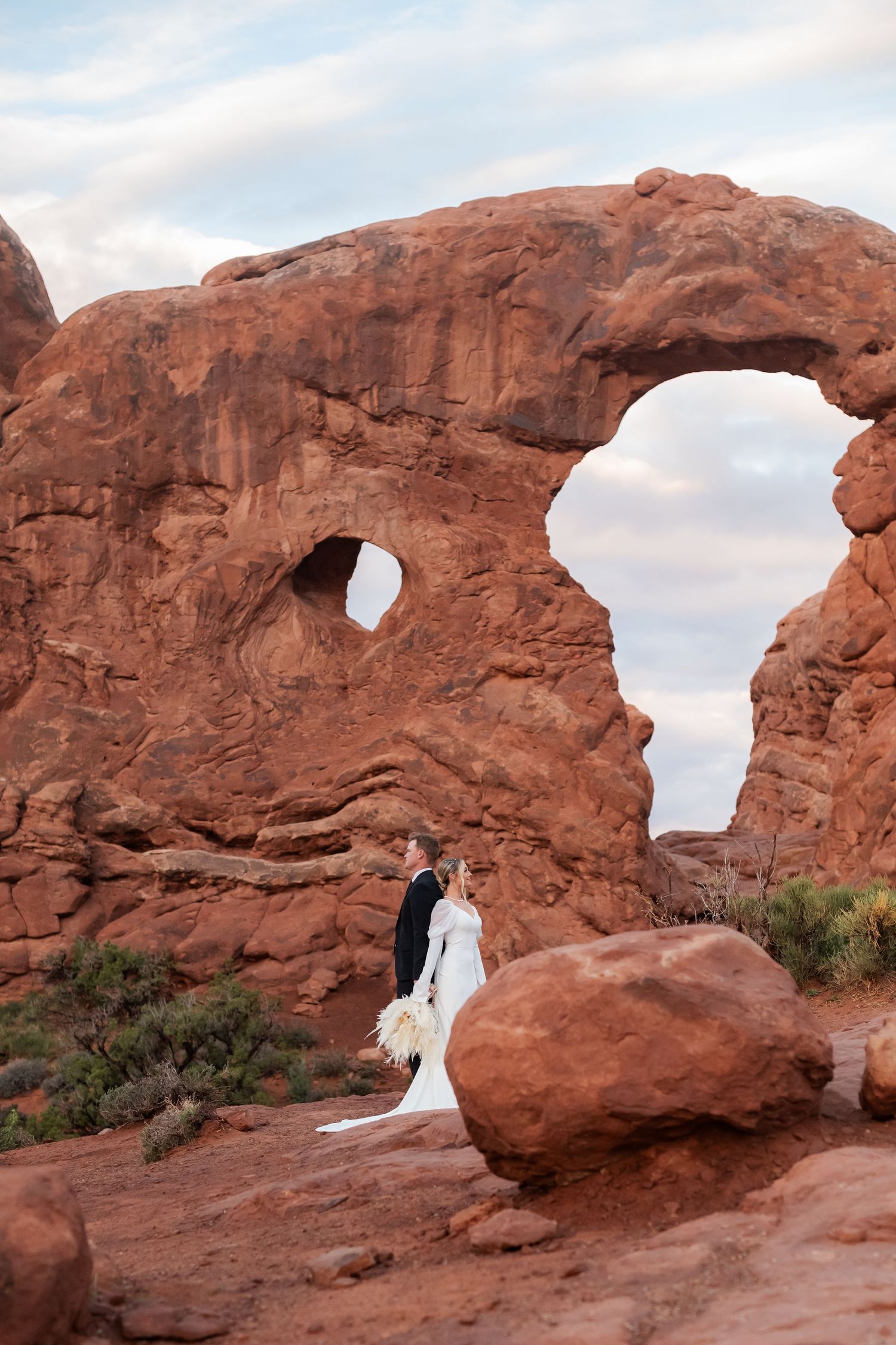 Wedding couple framed by natural rock arch formations in desert setting.