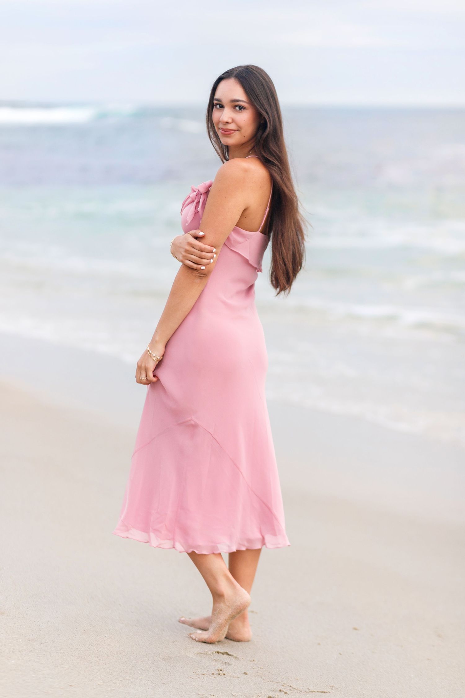 A person in a flowing pink sundress walks along a sandy beach with ocean waves in the background.
