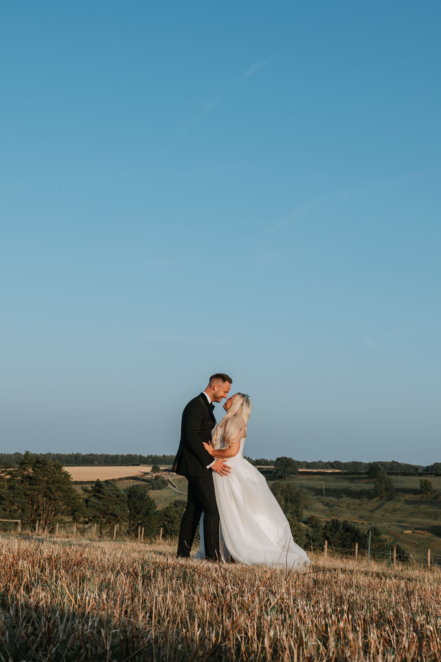 A couple in wedding attire embrace on a hilltop during golden hour with rolling countryside in the background. Kingscote Barn Wedding.