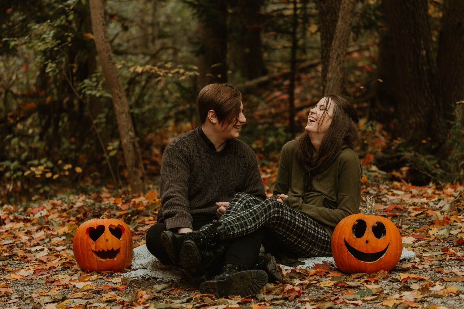 A couple sits on autumn leaves in the woods with two carved jack-o'-lantern pumpkins beside them.