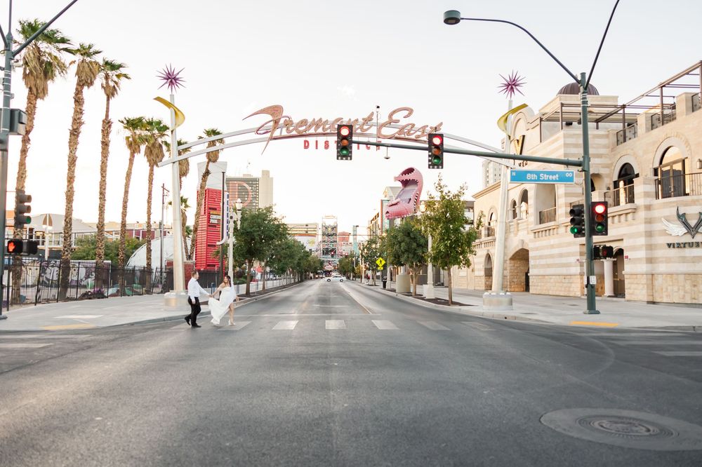 Palm trees line an urban street intersection with traffic lights and commercial buildings in a desert city setting.