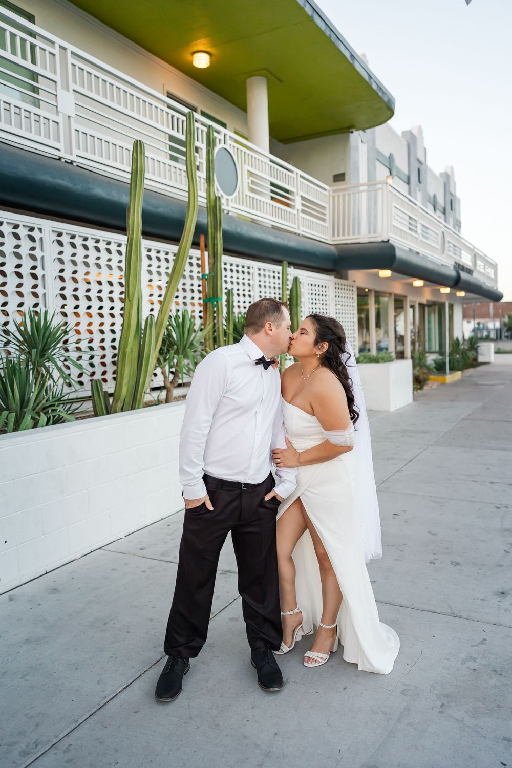 A romantic couple in wedding attire shares intimate moments along a retro motel exterior walkway.