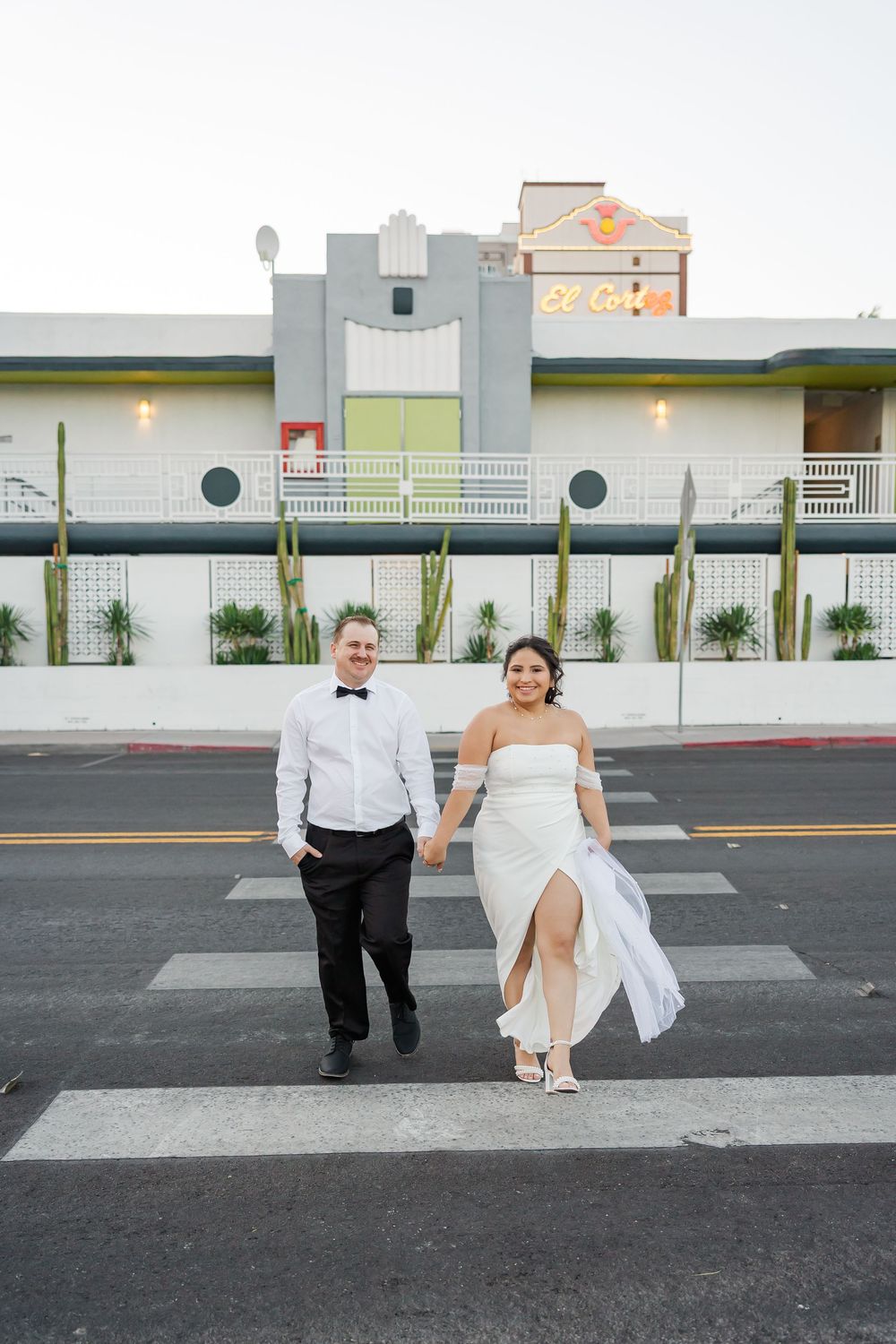 A couple in formal wedding attire walk together across a crosswalk in front of a modern building exterior.