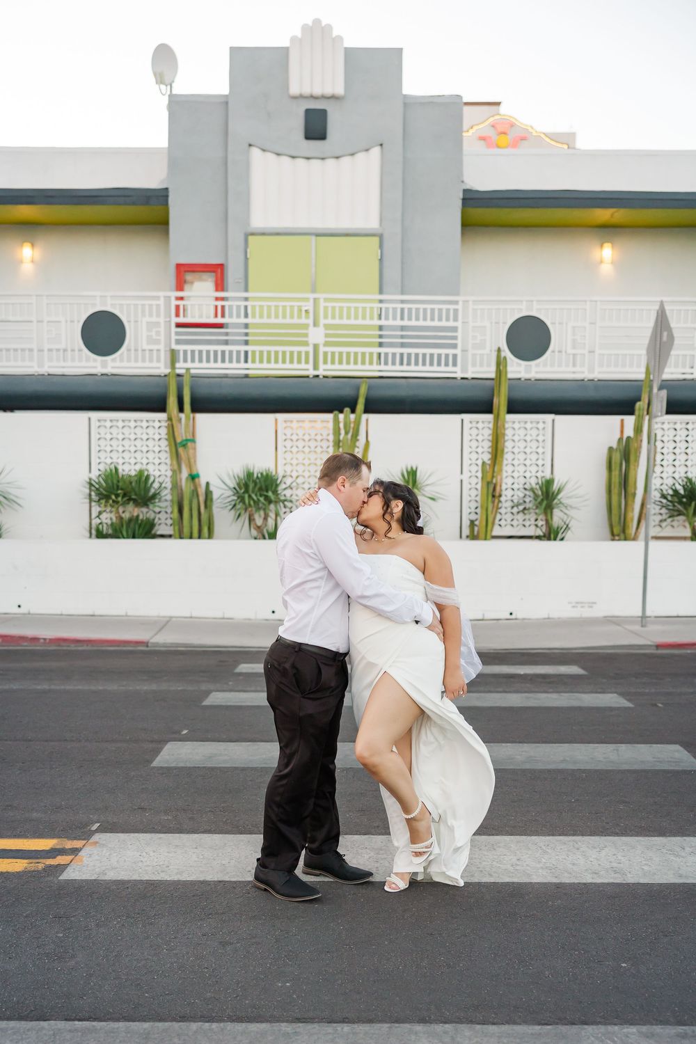 A couple in formal wedding attire embraces while kneeling on a crosswalk in front of a white building.