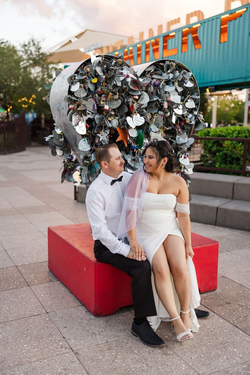 A couple in wedding attire sits on a red bench outdoors, sharing a romantic moment together.