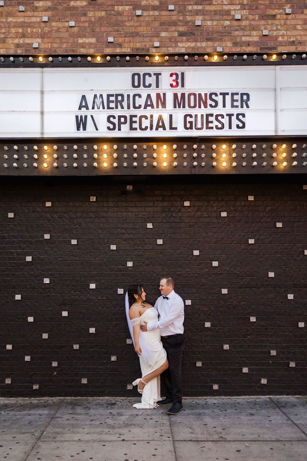A couple poses for wedding photos against a dark brick wall with a theater marquee in the background.