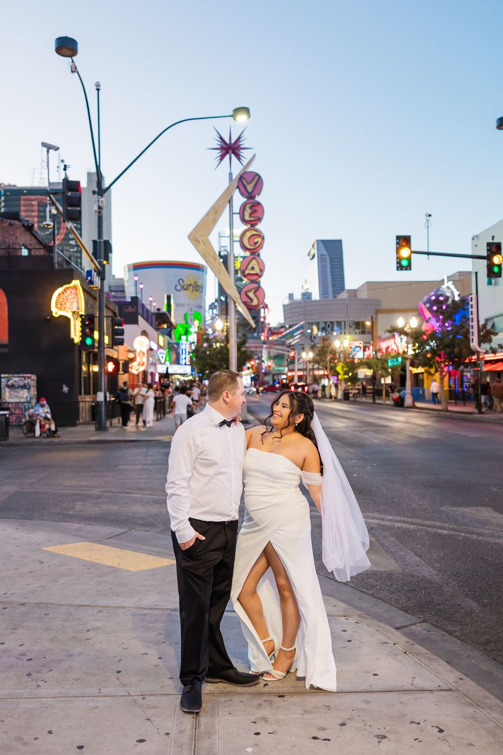 A couple poses for wedding photos on a city street with neon signs and lights in the background.