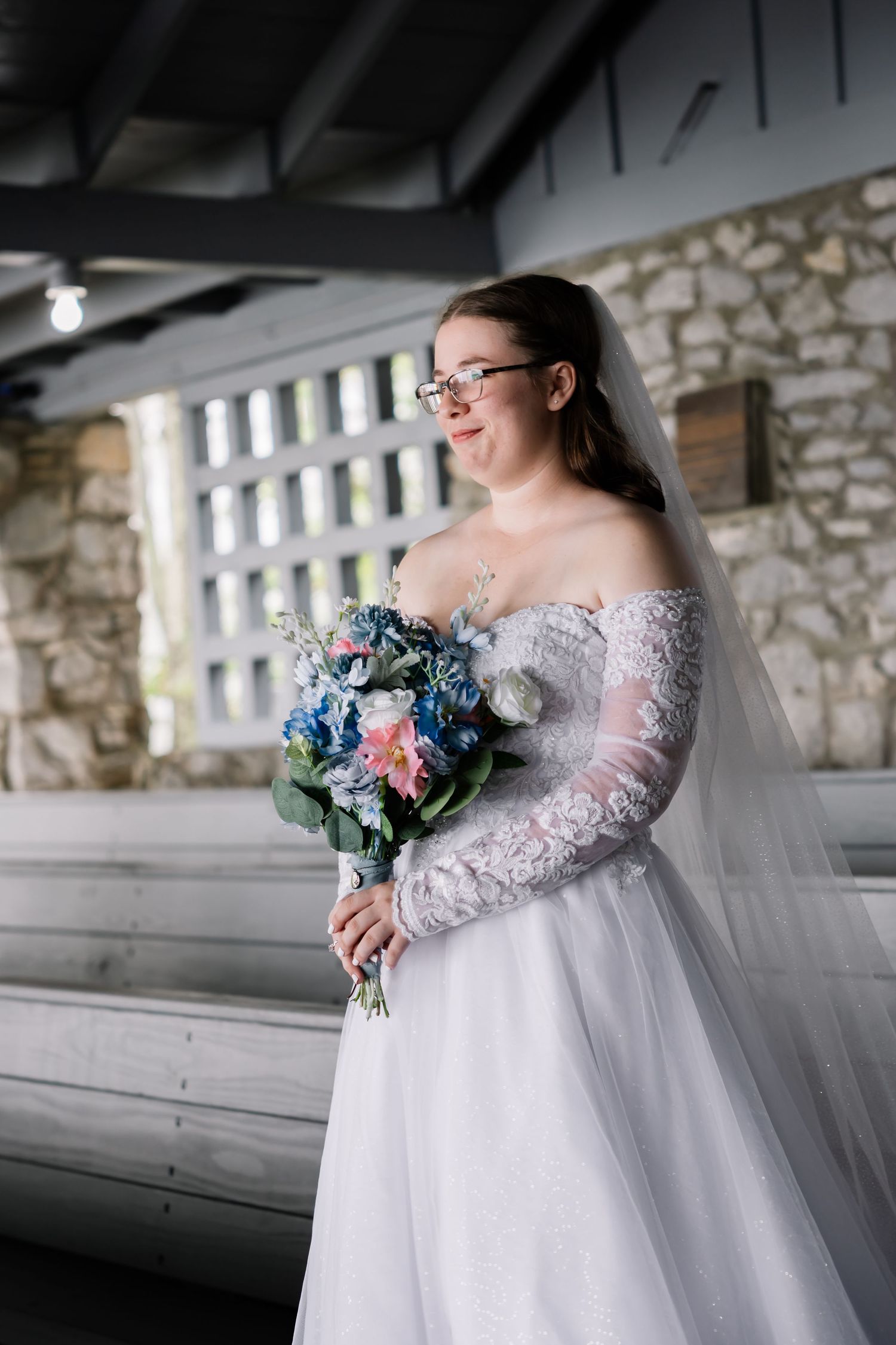 A bride in a lace wedding gown holds a blue and pink bouquet while posing in a rustic stone building.