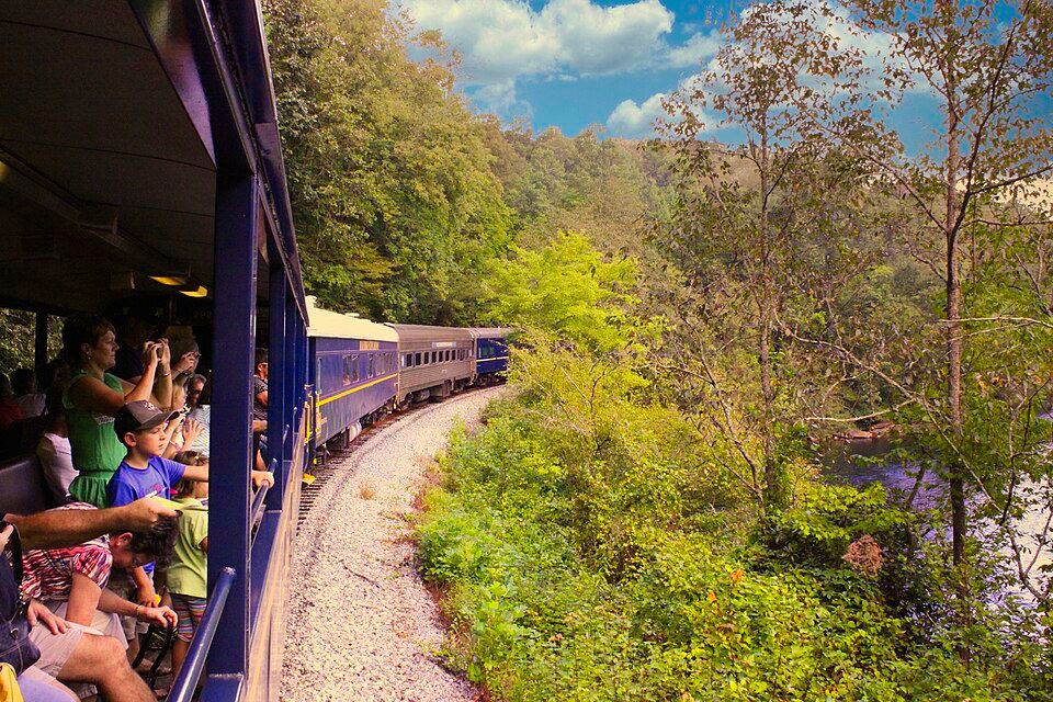 Passengers lean from open windows of a blue train winding through a lush green river gorge.